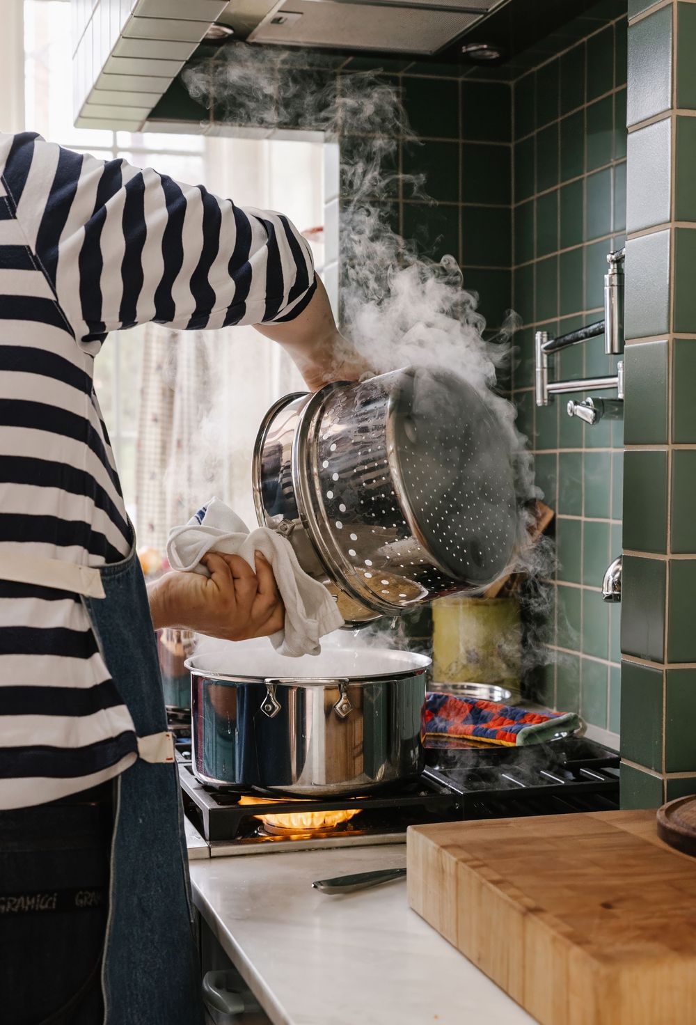 Person lifting steamer basket over a pot on a stove in a green-tiled kitchen.