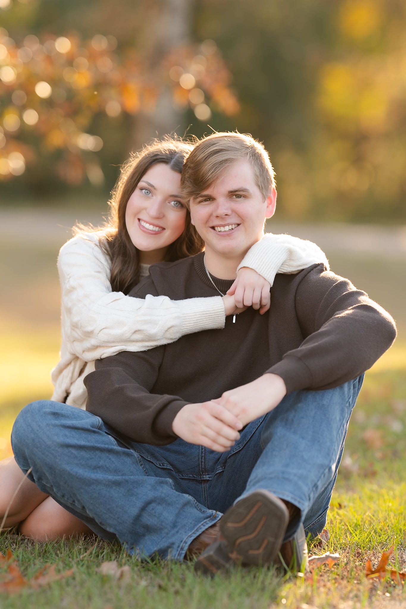 A couple sits together on grass during golden hour with autumn bokeh lights in the background.