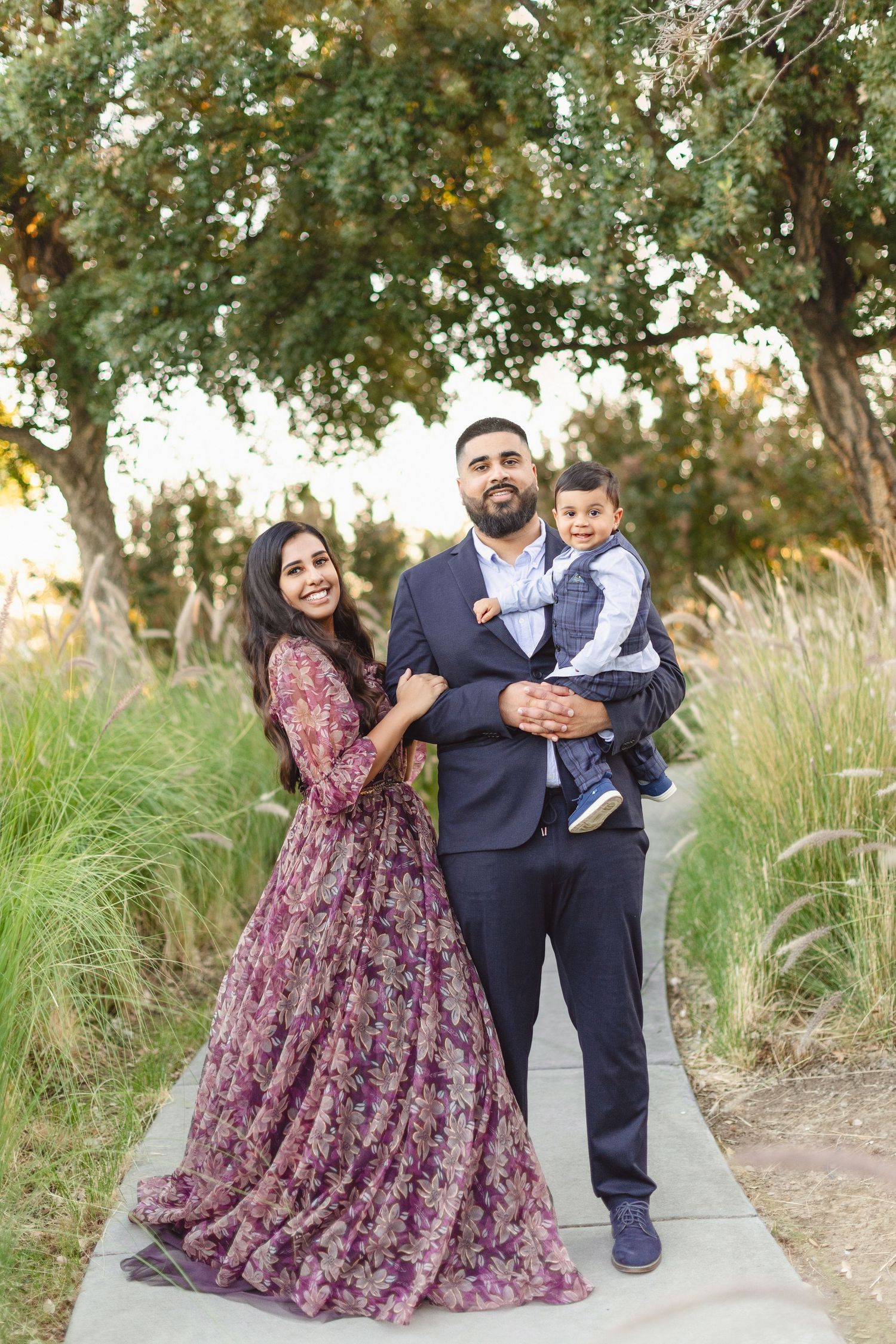 Family of three in formal attire posing on pathway with trees and tall grass in background.