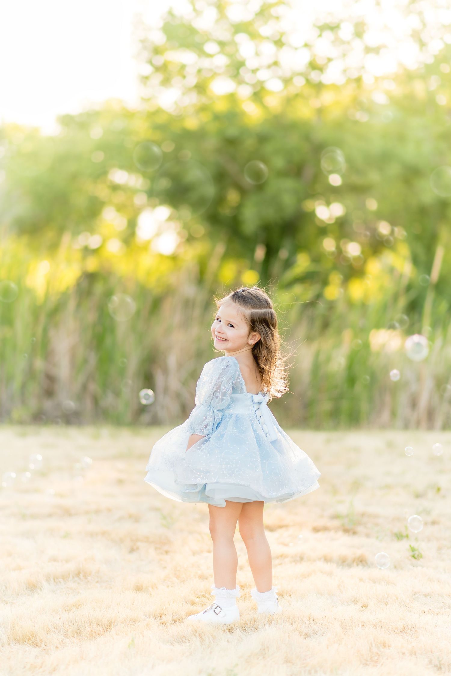 Young child in light blue dress twirling among bubbles in golden sunset field.