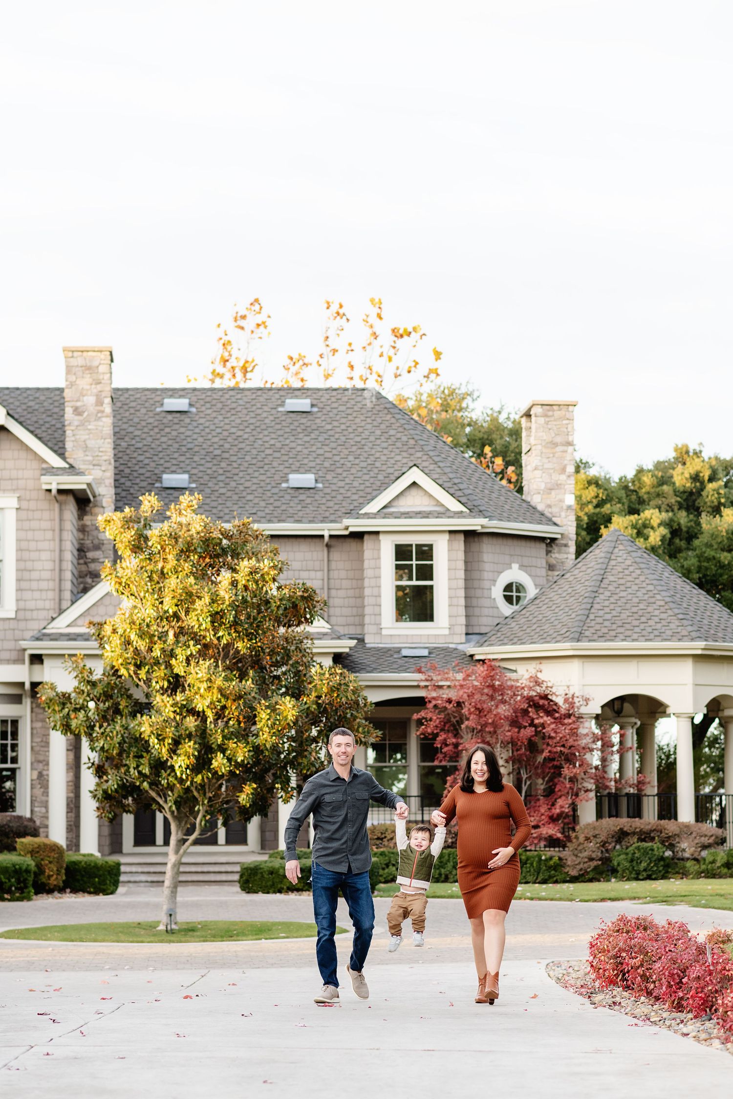 Couple running playfully together on driveway in front of luxurious suburban home with fall foliage.