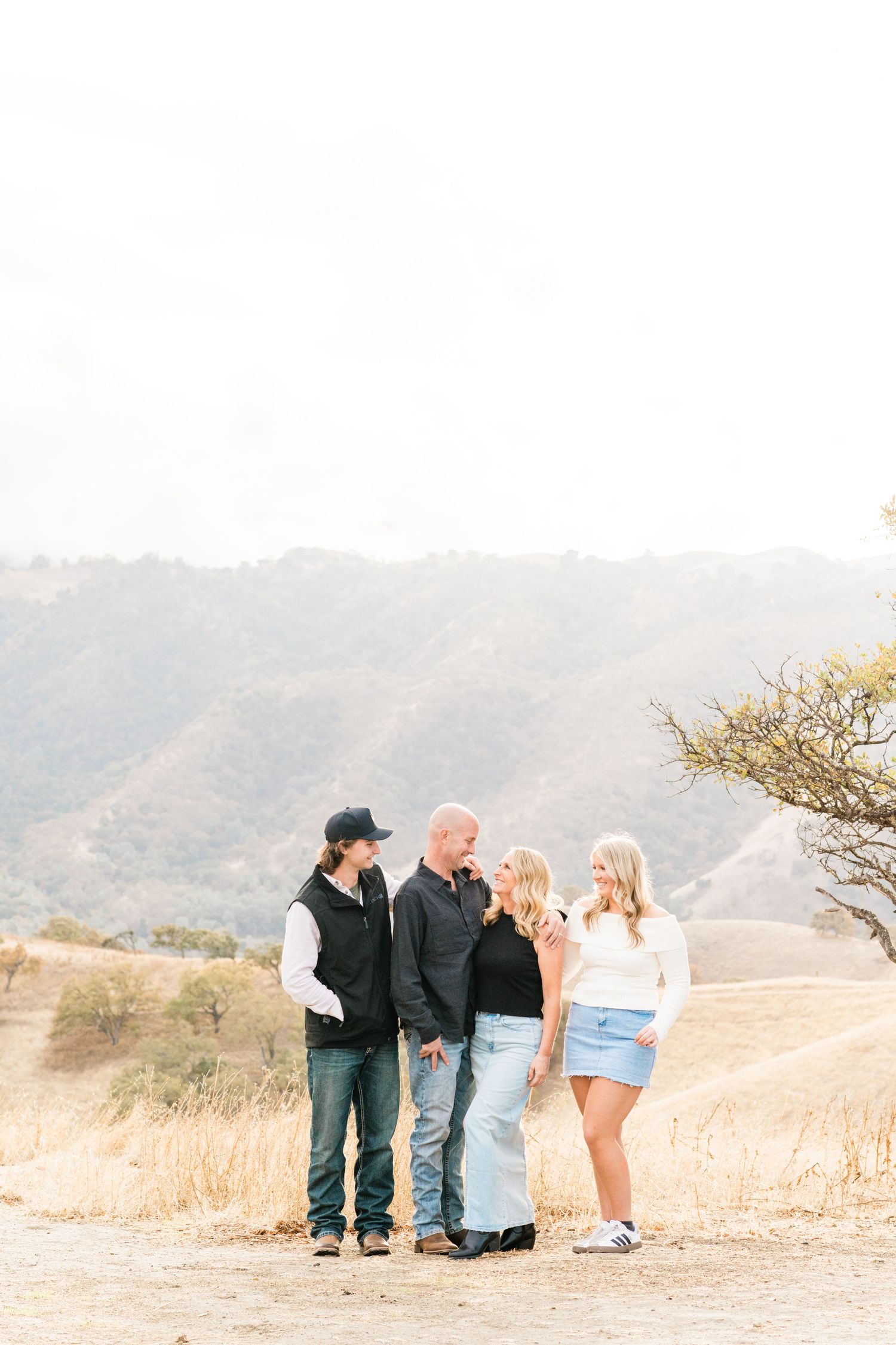 Family of four smiling and embracing while standing on a scenic hillside trail during golden hour.