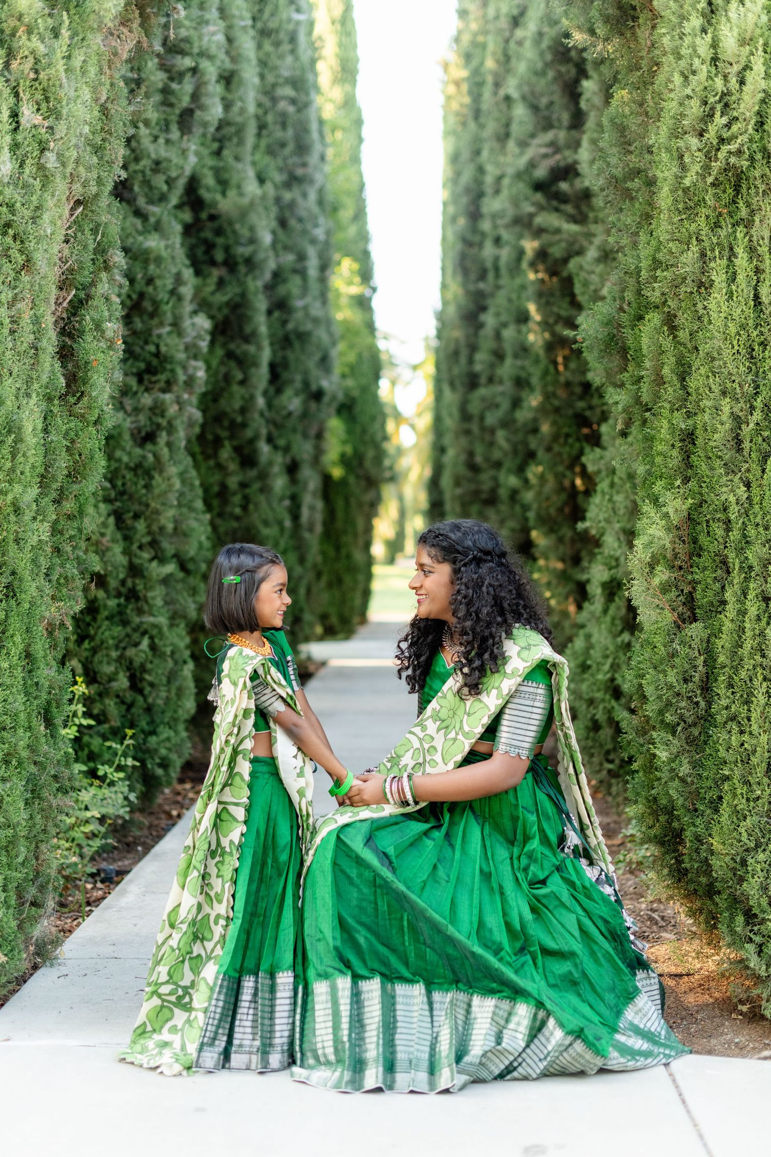 Two people in matching green saris pose together on pathway lined with towering cypress trees.