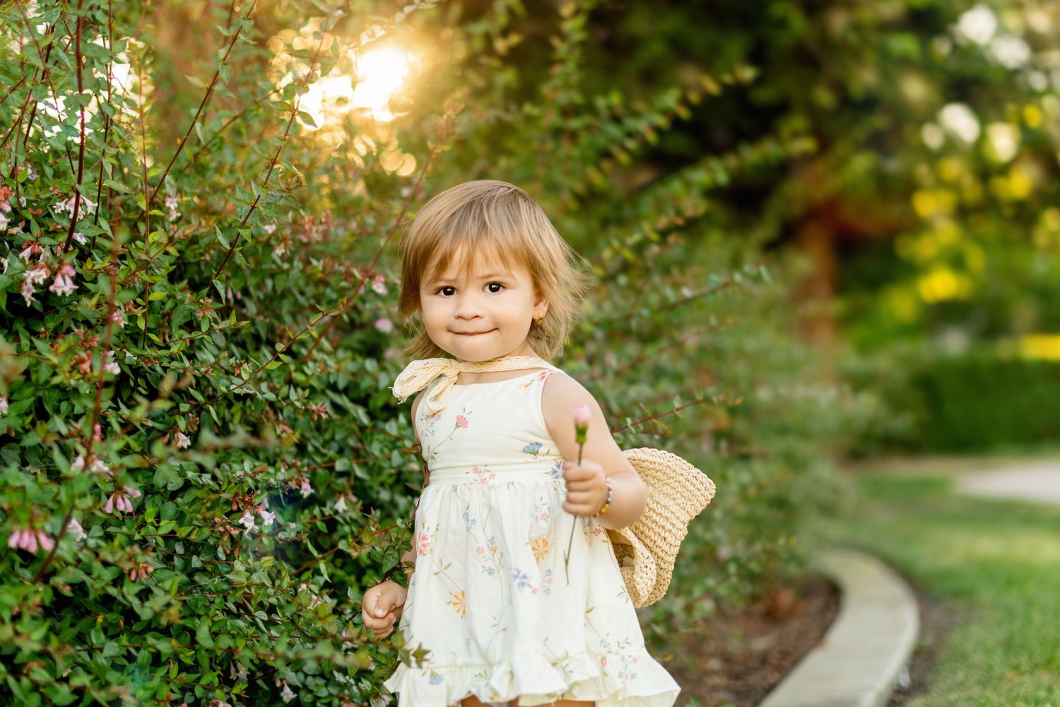 Young child in white dress smiling in sunny garden setting.