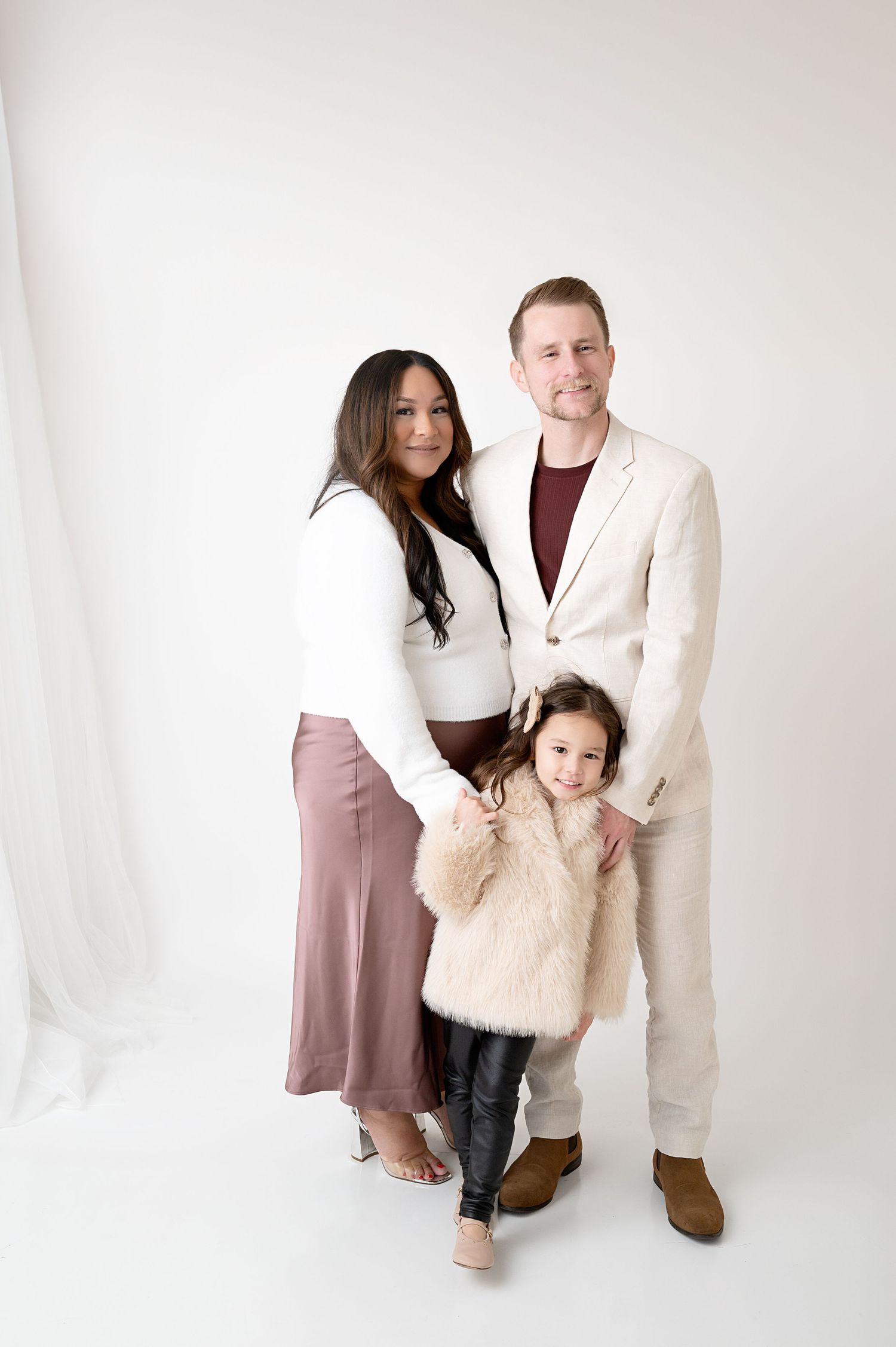 A stylish family portrait against a white backdrop featuring coordinated winter outfits in neutral tones.