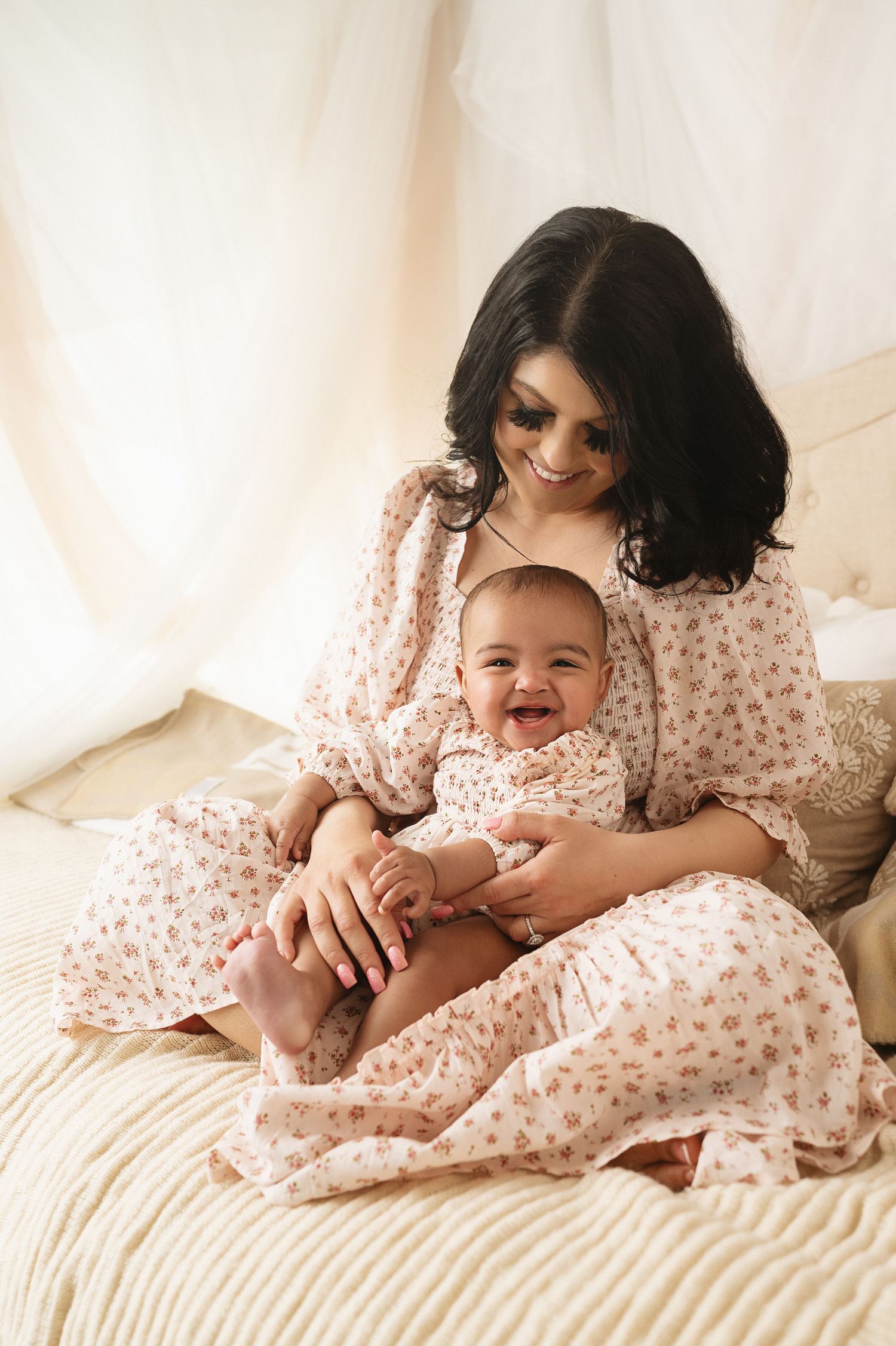Mother and baby in matching floral dresses sharing tender moment on white bedding.
