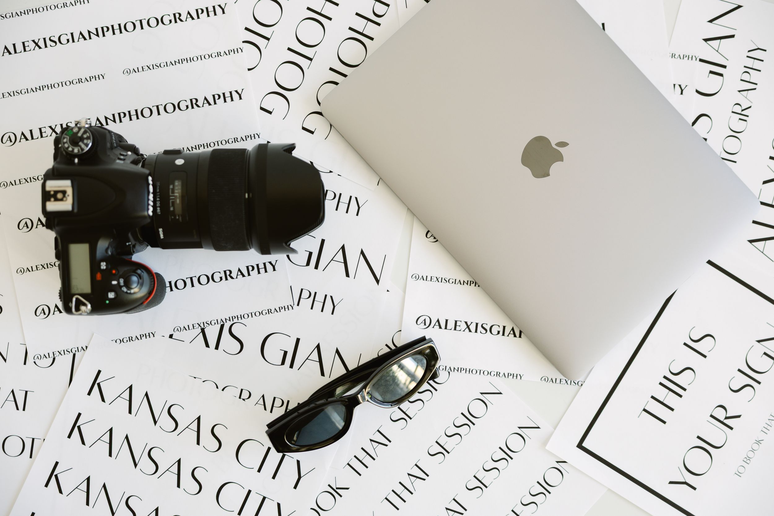 Flat lay of camera, MacBook, sunglasses, and branded photography prints on white surface during a branding session in natural light studio.