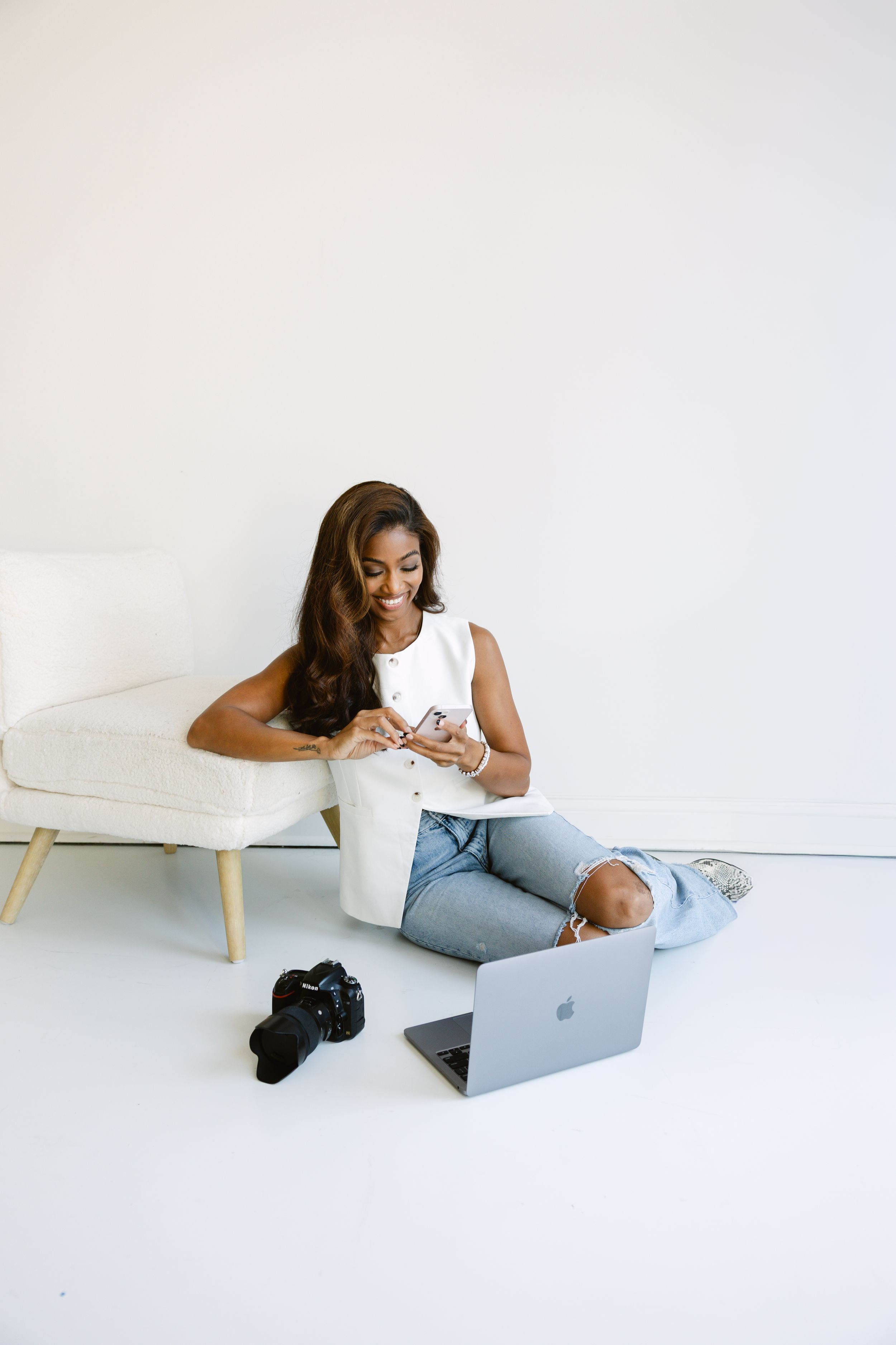 A photographer sitting on floor with phone in hand and laptop and camera on floor during a branding session at a natural light studio.