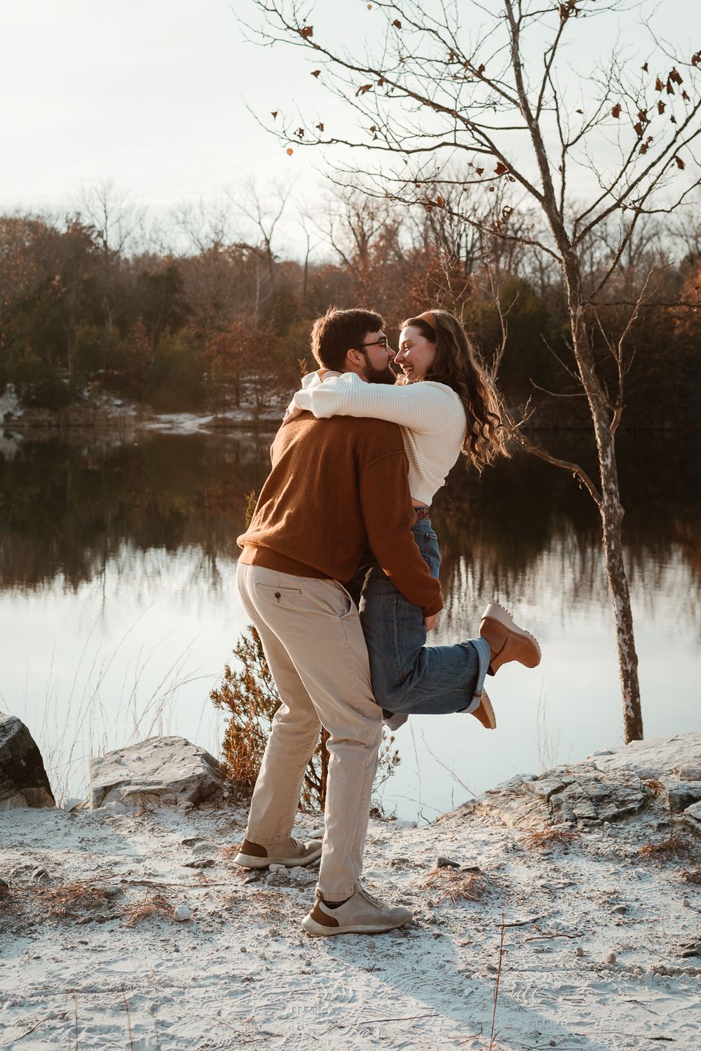 A romantic couple embracing by a lake during sunset with autumn trees in the background.