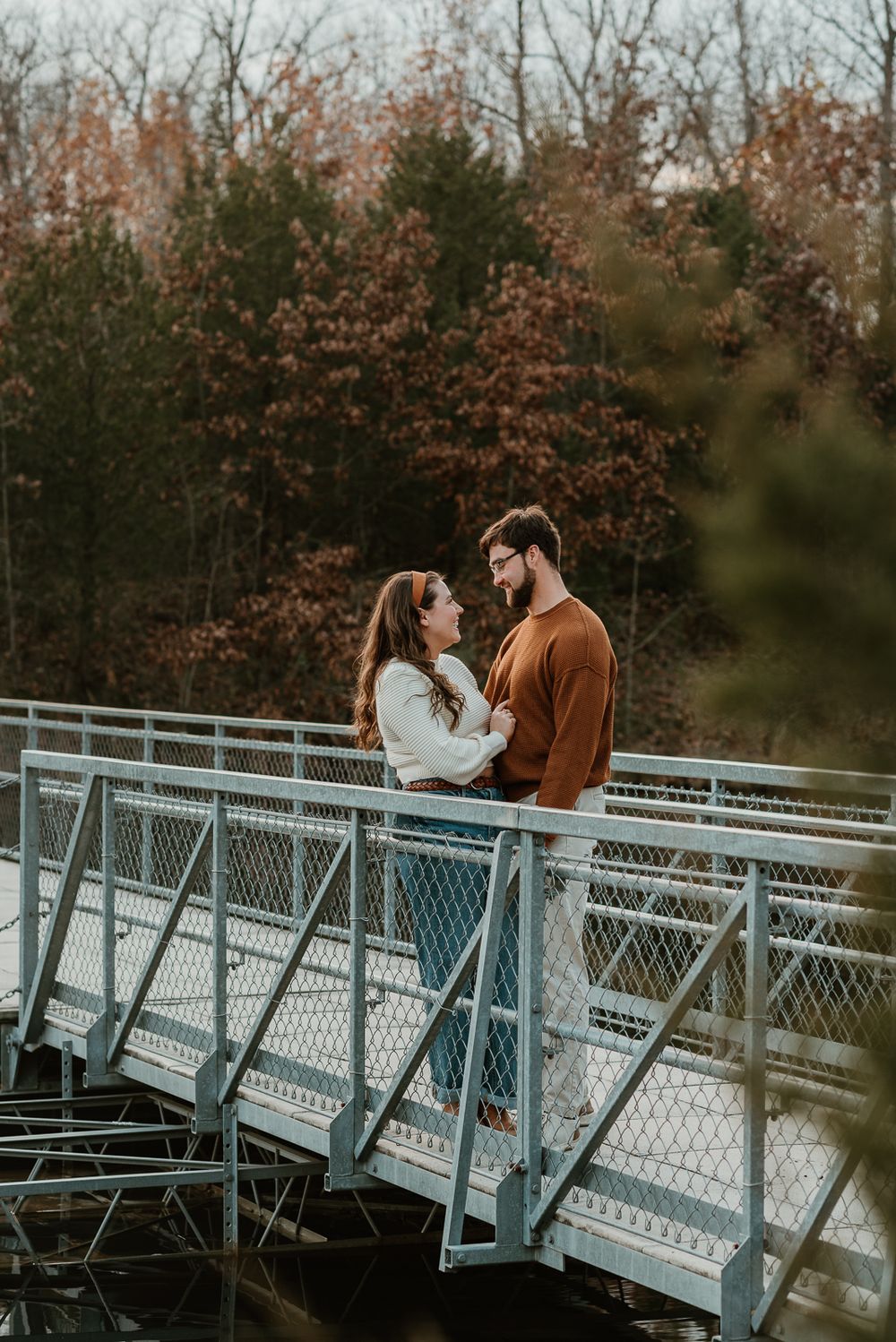 A couple sharing a romantic moment on a metal bridge with fall foliage in the background.