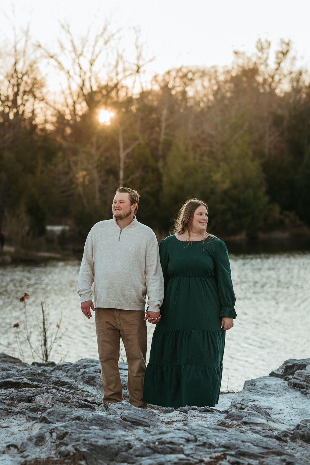 A couple stands together on rocky terrain by a lake during golden hour.