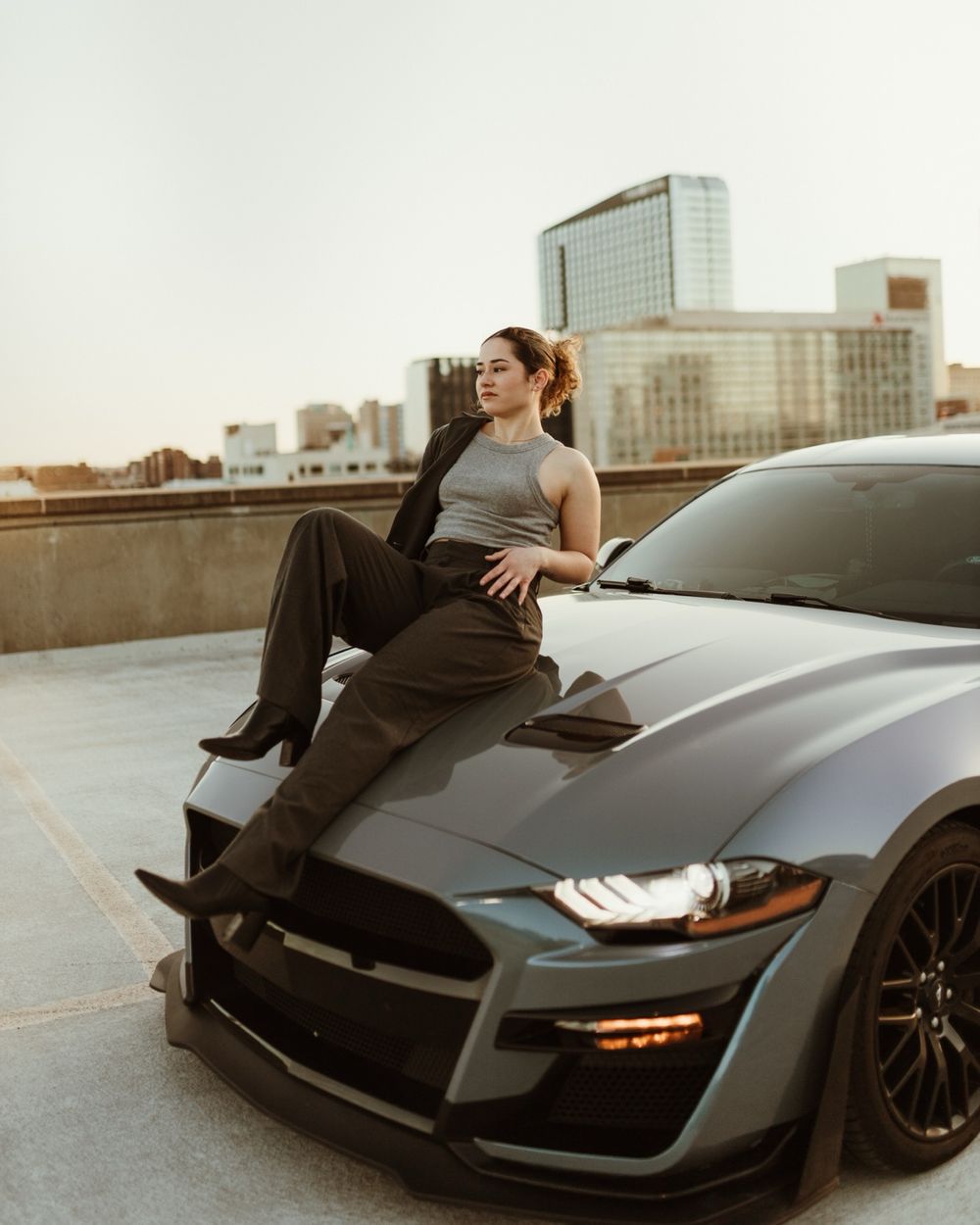 a woman sitting on top of a black sports car, on top of a parking garage that overlooks a city skyline.