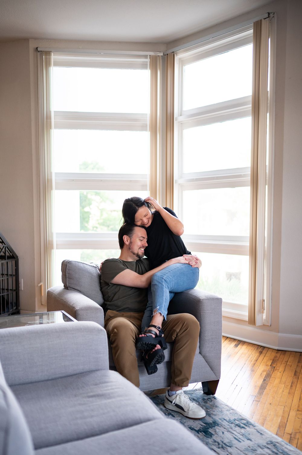 Two people sharing an intimate moment while relaxing on a gray couch near bright windows.