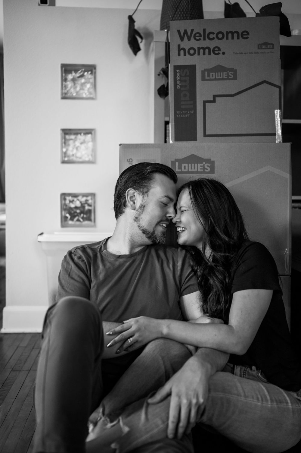 A couple embracing near moving boxes with a 'Welcome Home' sign visible in the background.