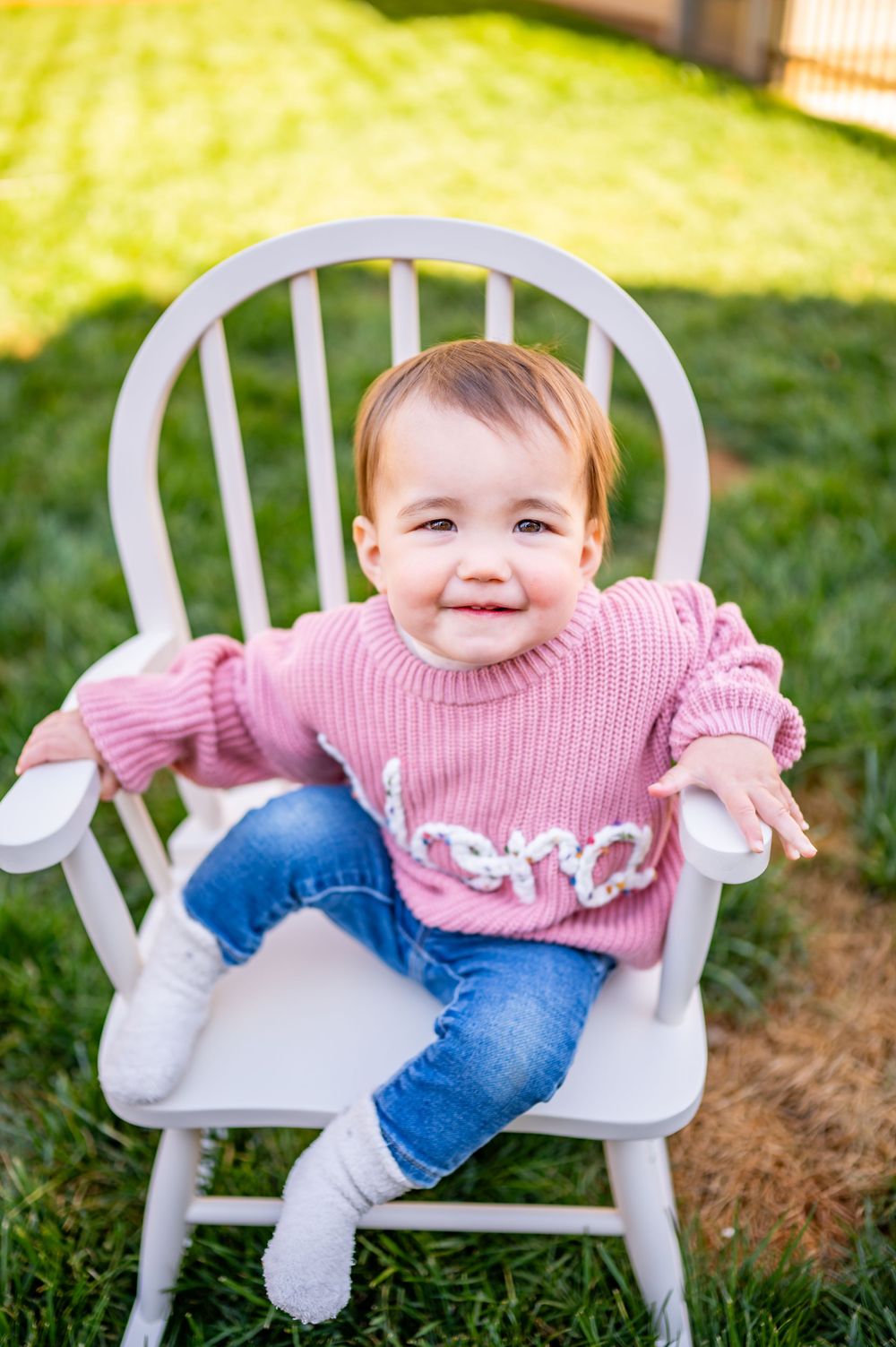 Happy baby wearing pink knit sweater and jeans smiles while sitting in white chair outdoors.