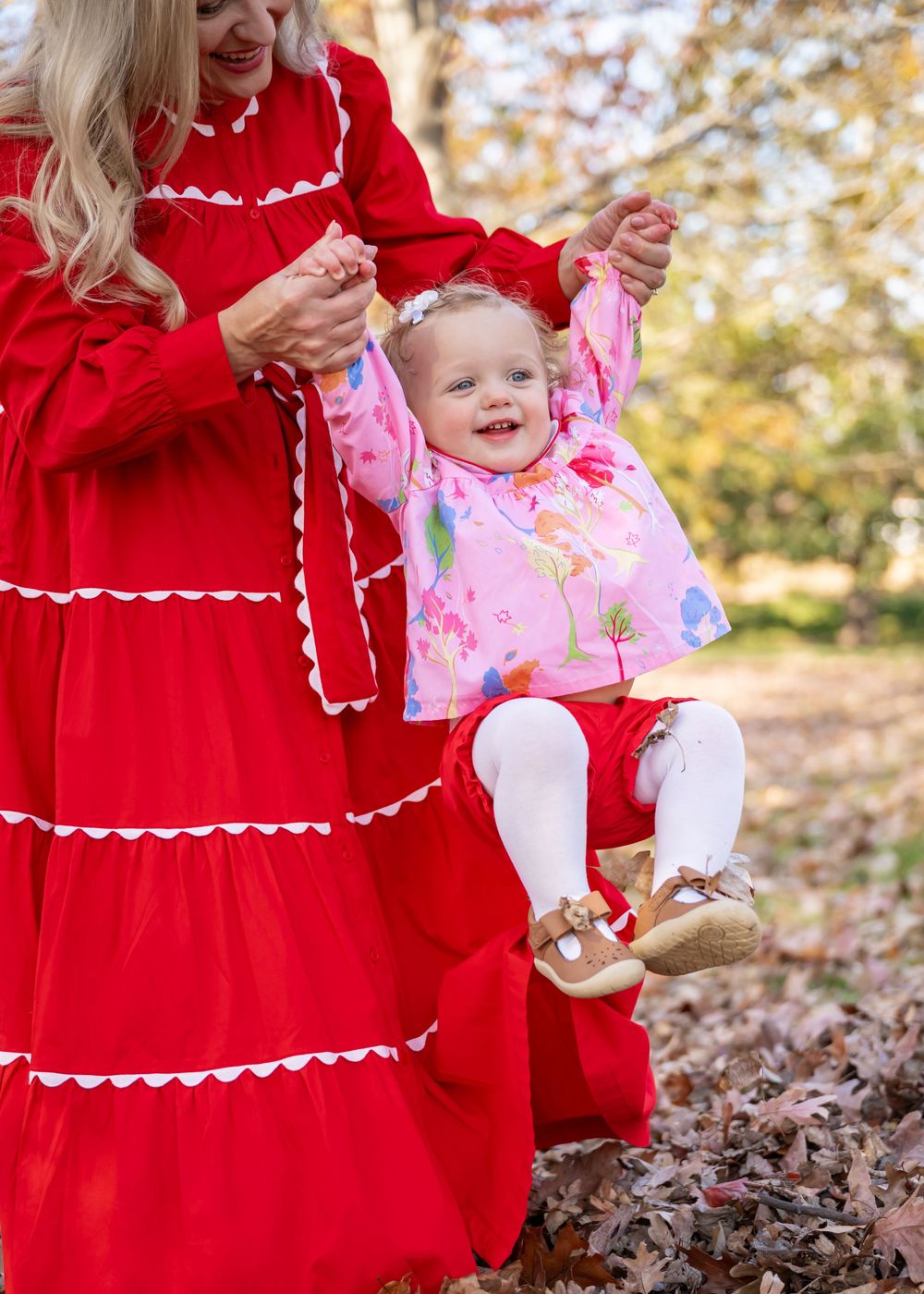 A mother and daughter in matching red dresses sit together in fallen autumn leaves during an outdoor photo session.