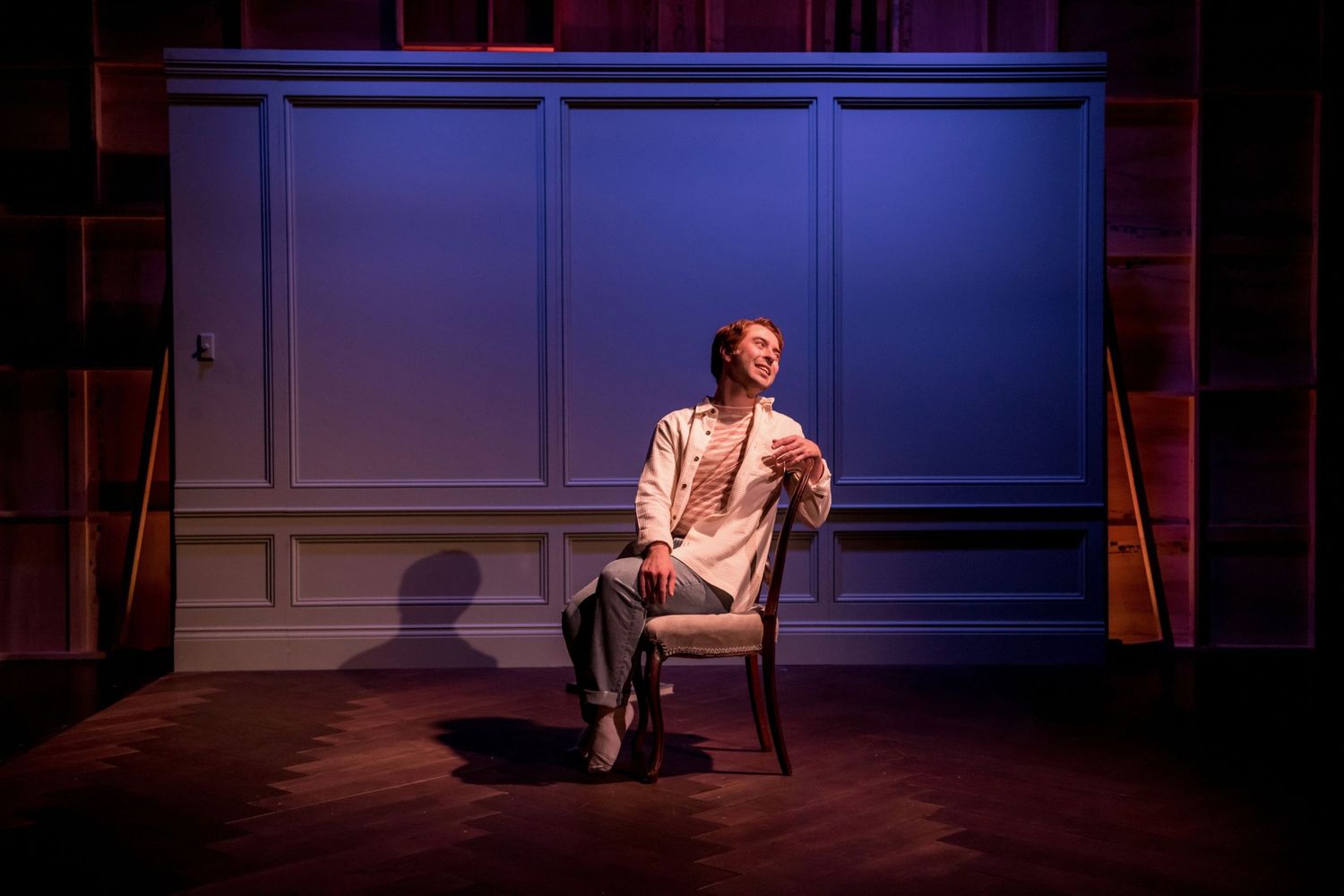 Solo performer seated in chair against blue wall creates intimate theatrical moment.