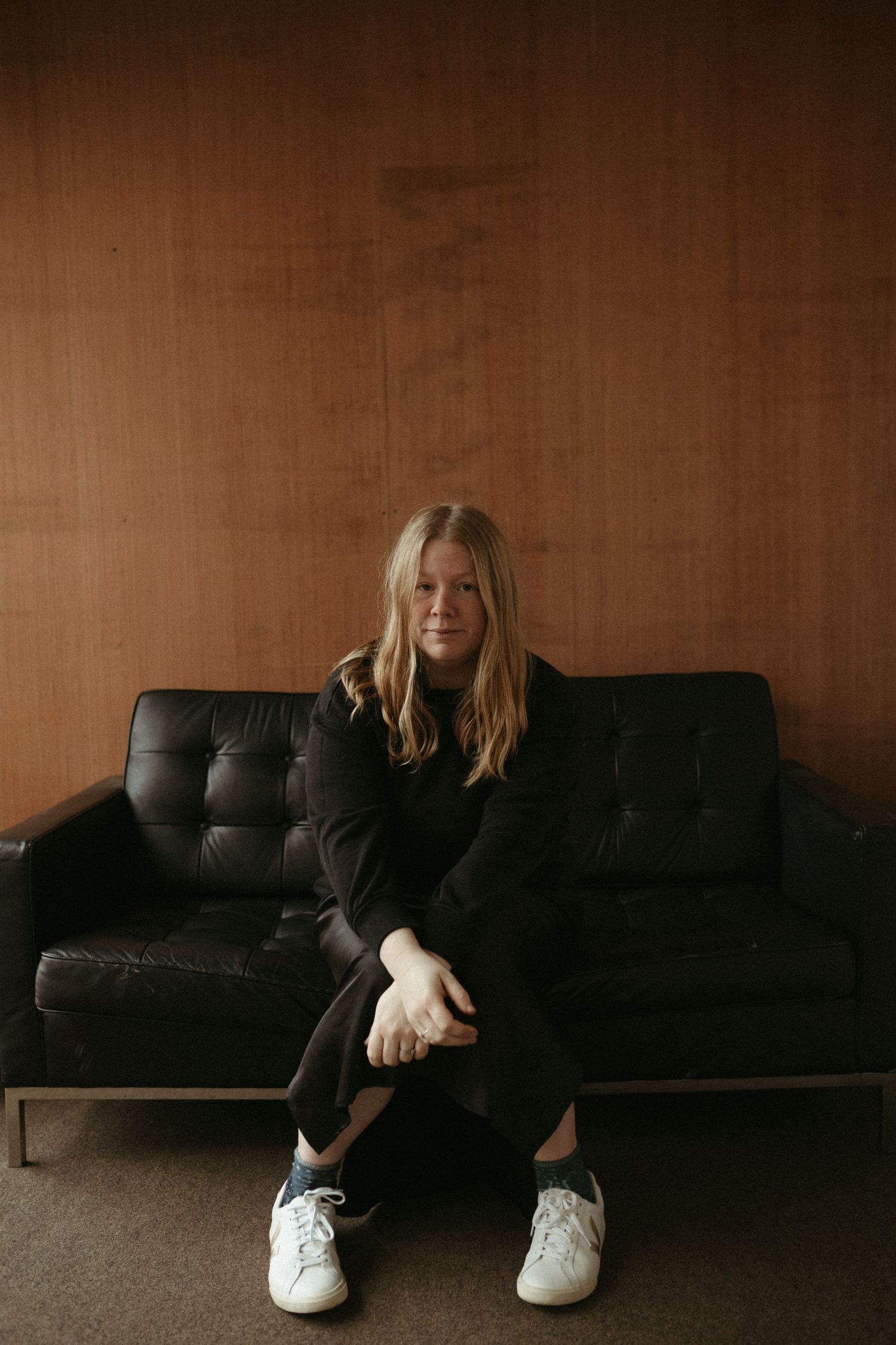 A person in all black attire sits casually on a dark leather couch against a brown wall wearing white sneakers.