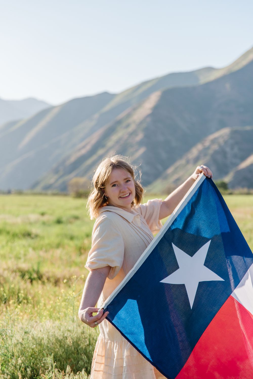 Person in cream dress holding Texas state flag against mountainous backdrop.