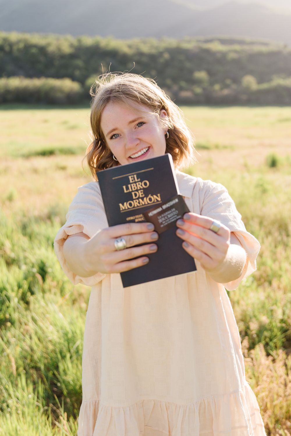 Person in white dress holding The Book of Mormon in a sunny field with mountains in background.