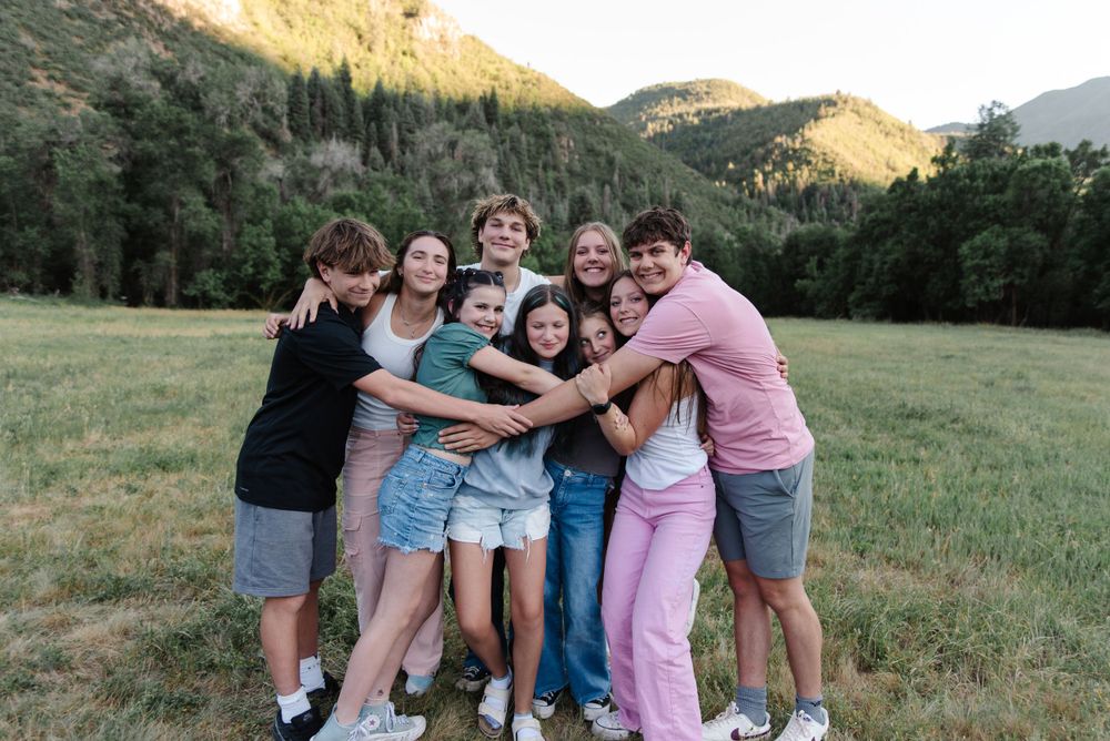 Friends sharing group hug in grassy field with mountain backdrop at sunset.