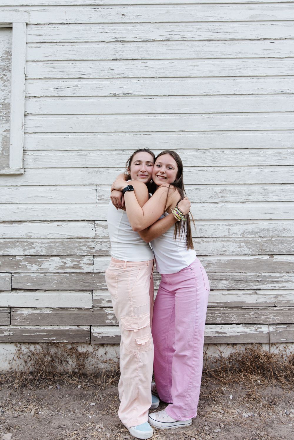 Two people in white tops and pink pants hugging against weathered white wooden wall.