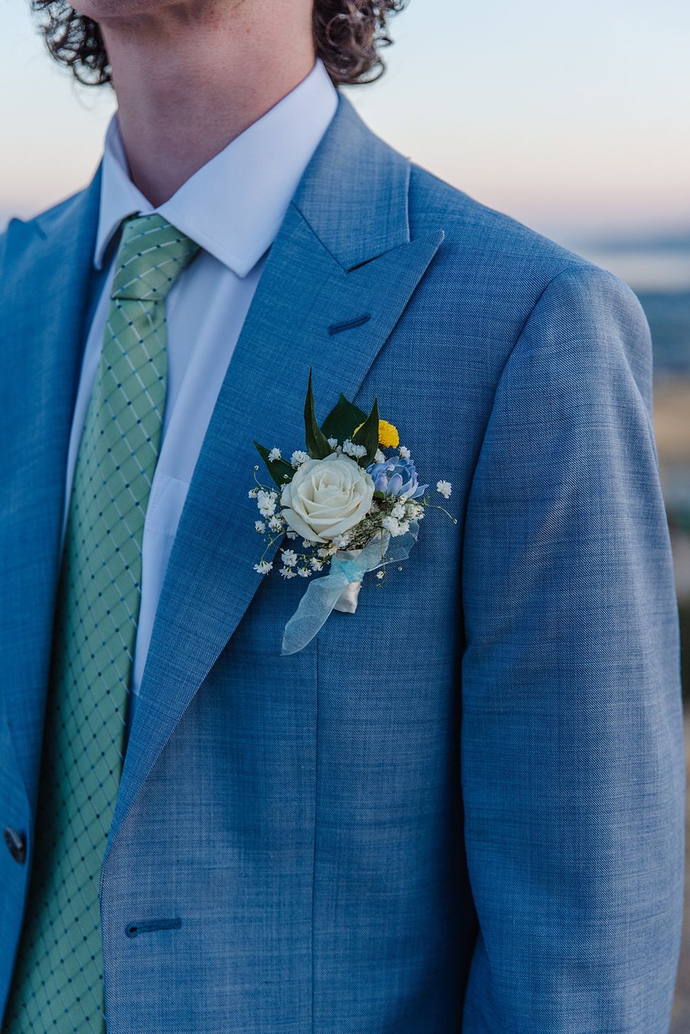 Close up of light blue suit jacket with white rose boutonniere and mint green tie.