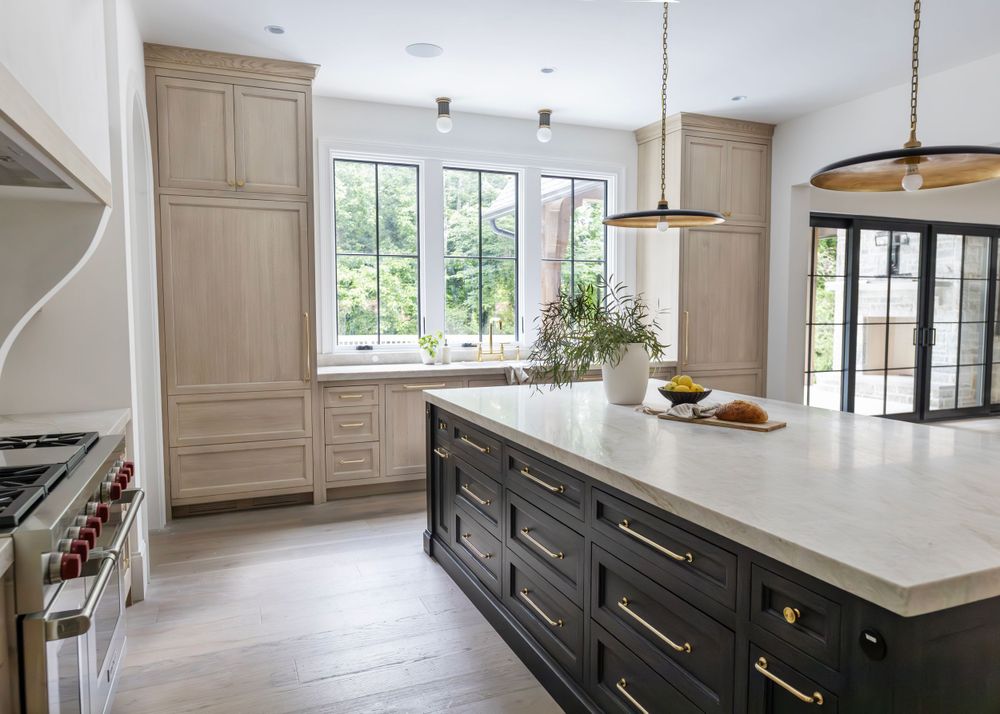 Modern kitchen with light cabinets, dark island, and large windows overlooking greenery.