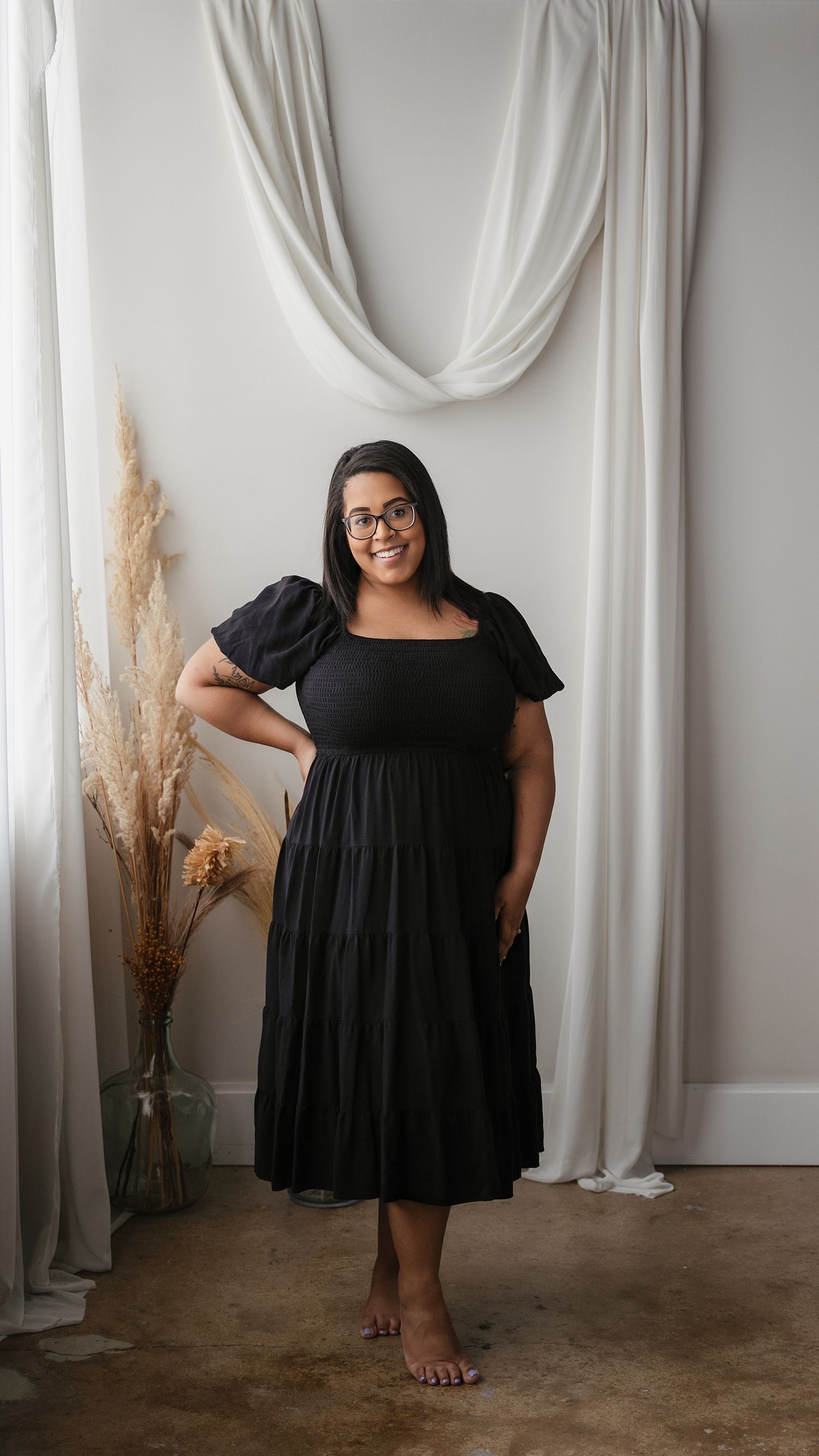 Woman in black midi dress poses against white draped backdrop with pampas grass decor.