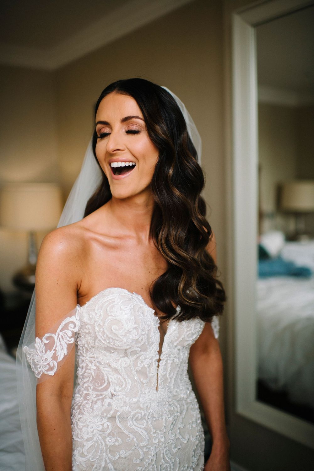 Beautiful bride wearing a strapless lace wedding dress with wavy dark hair laughs joyfully during wedding preparations.
