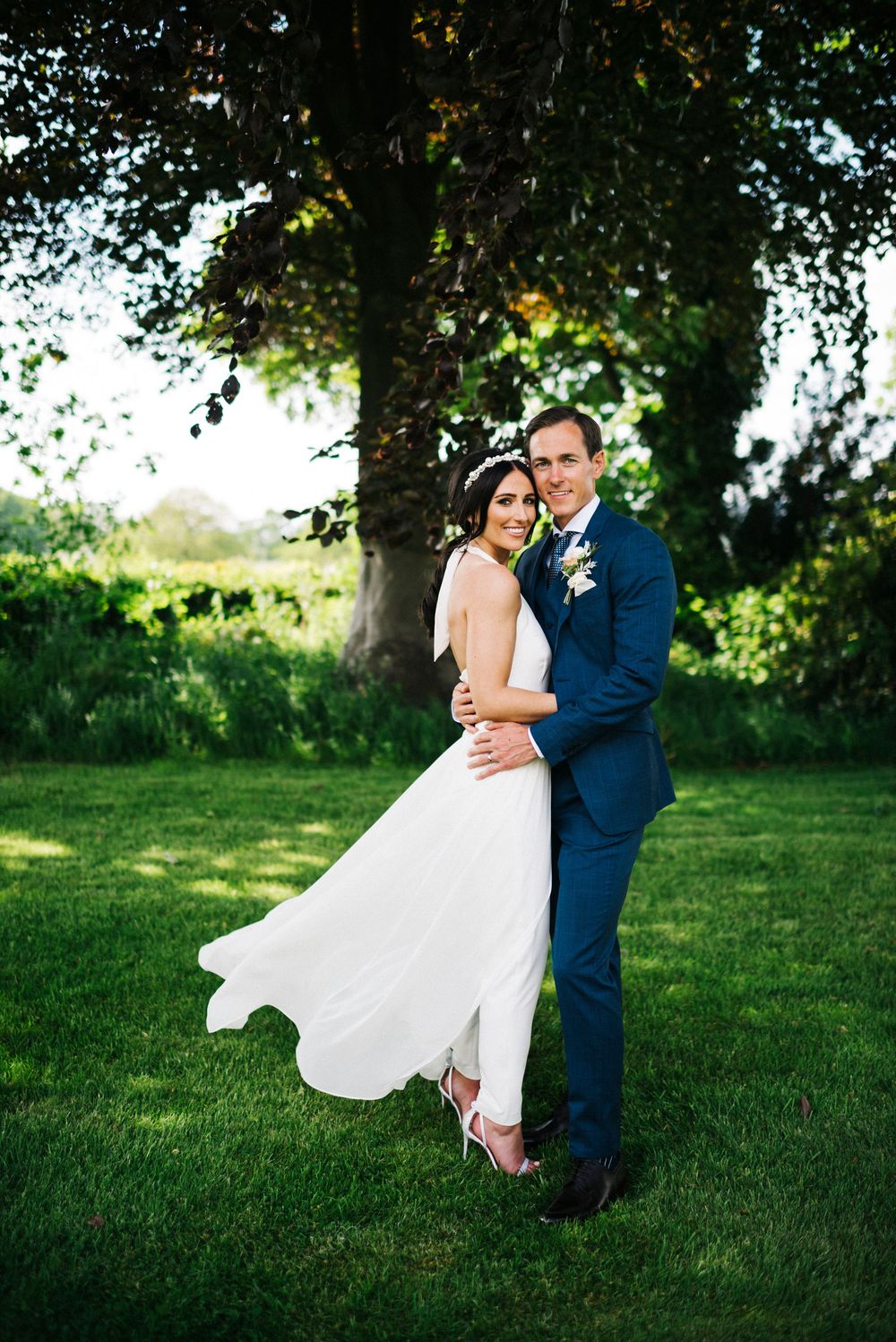 A couple in wedding attire dances and twirls together on a lush green lawn beneath shady trees.