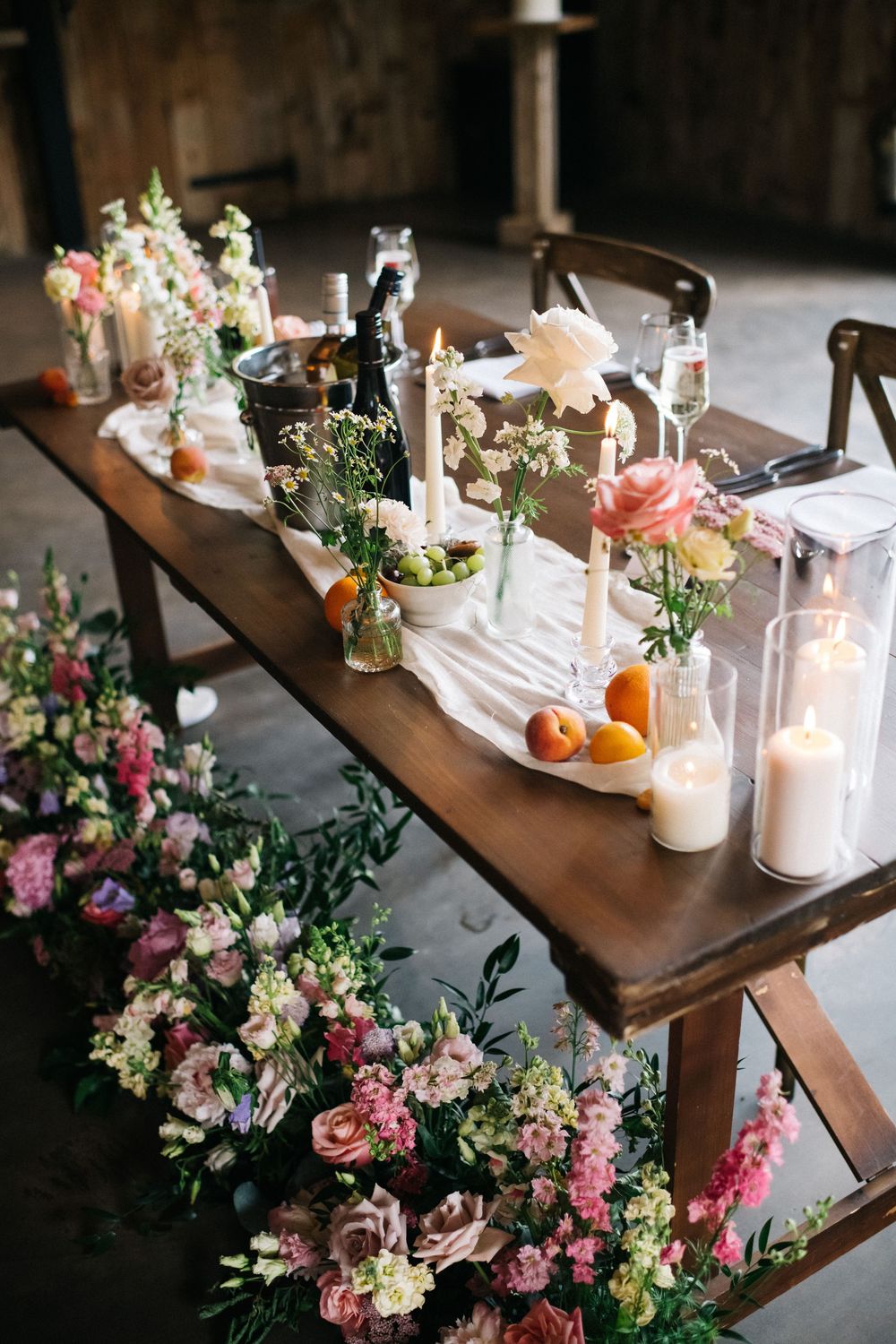 Long wooden dining table decorated with pink floral arrangements, white candles and rustic table settings for a romantic event.