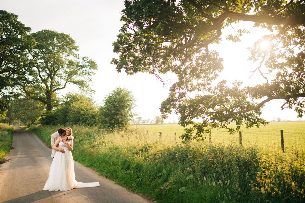 A couple embraces in a golden sunlit field during sunset with trees silhouetted in the background.