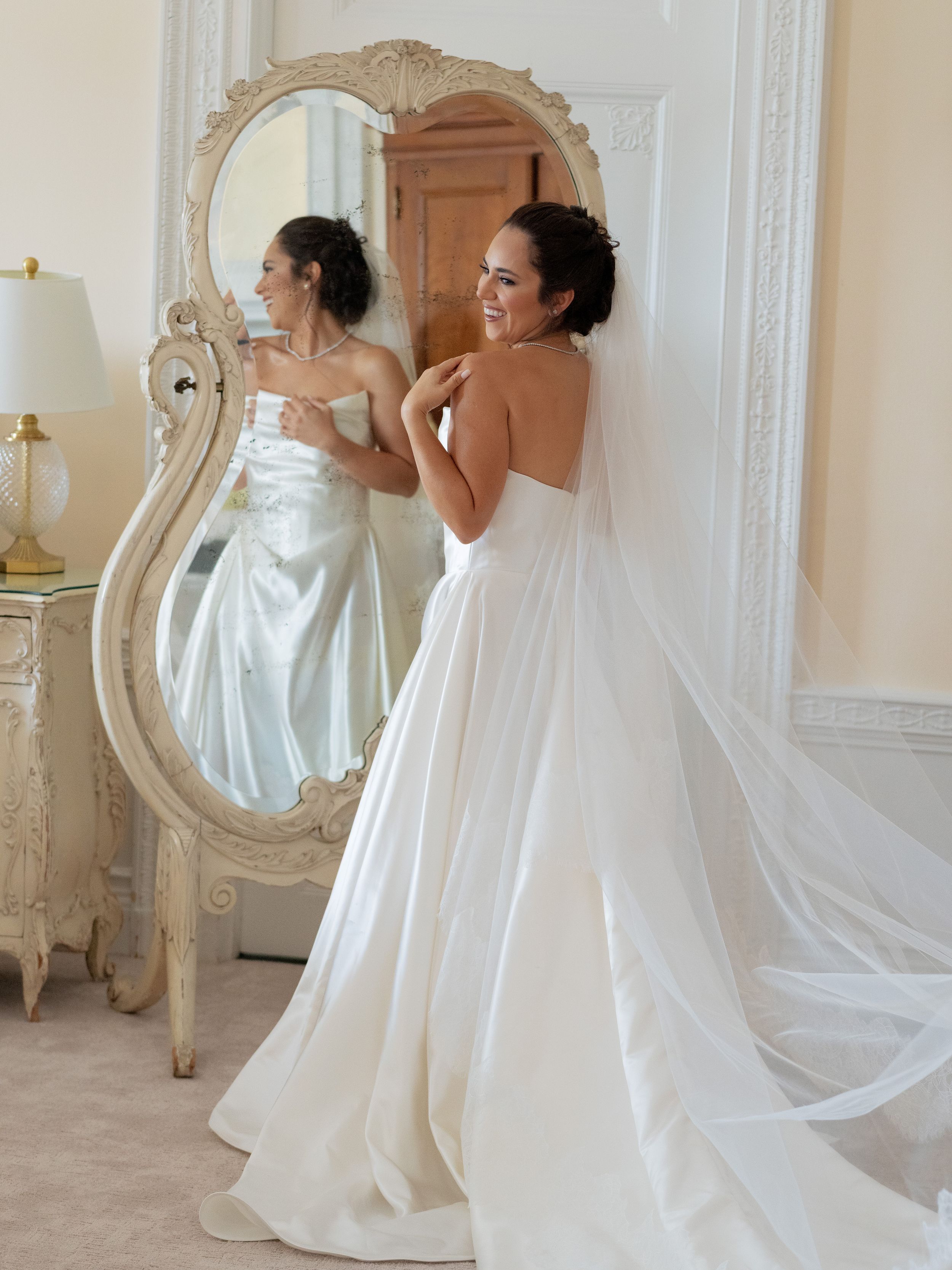 Elegant white wedding gown flowing in front of ornate vintage mirror.