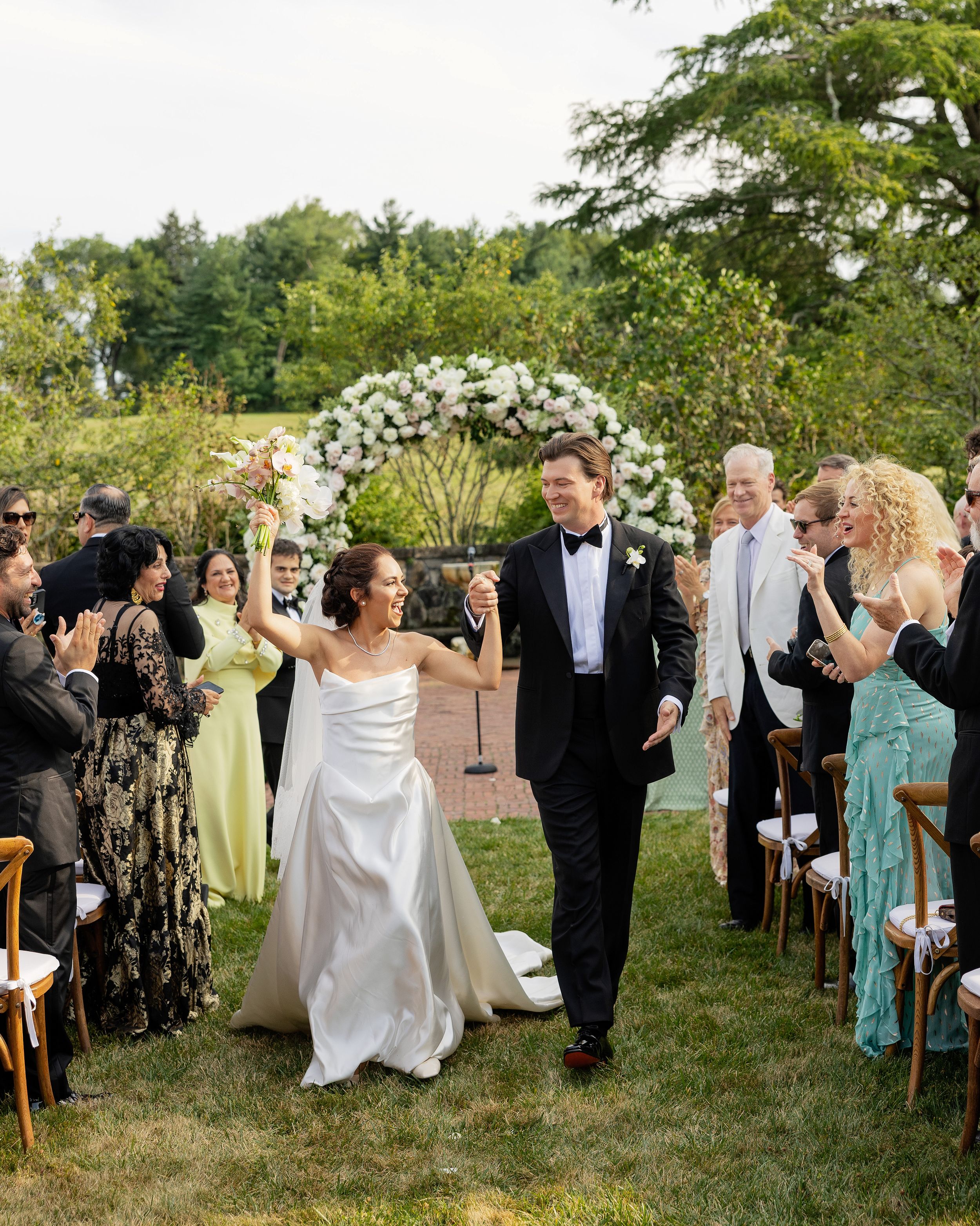 Outdoor wedding ceremony with guests in formal attire and turquoise dresses under trees.