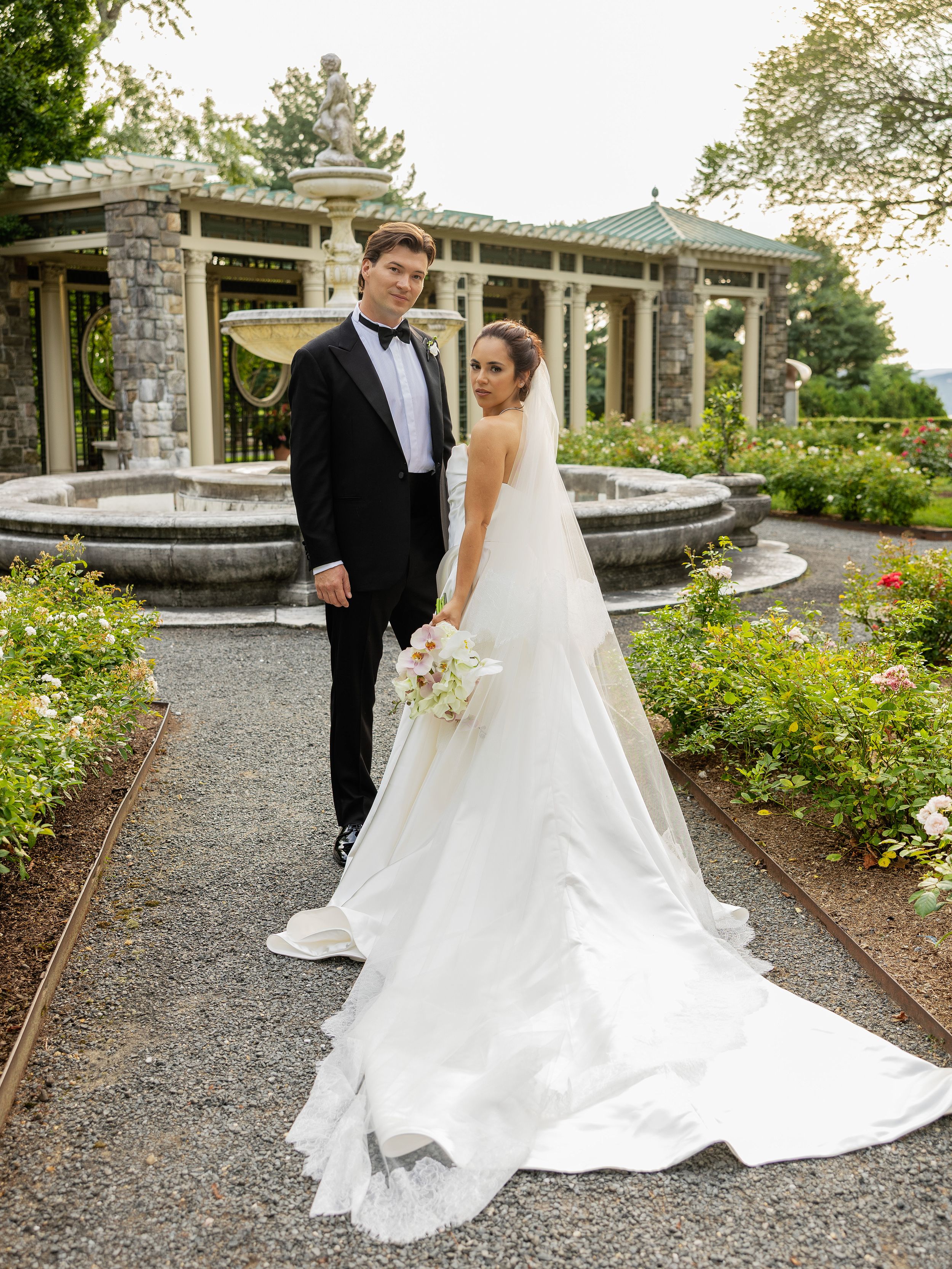 A wedding couple walking on a gravel path surrounded by lush gardens and stone architecture.