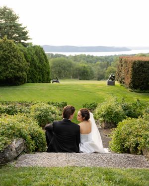 A couple sits on a stone wall overlooking a scenic water view on their wedding day.