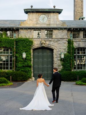 A couple walks along a beautiful stone building with ivy-covered walls and large wooden doors.