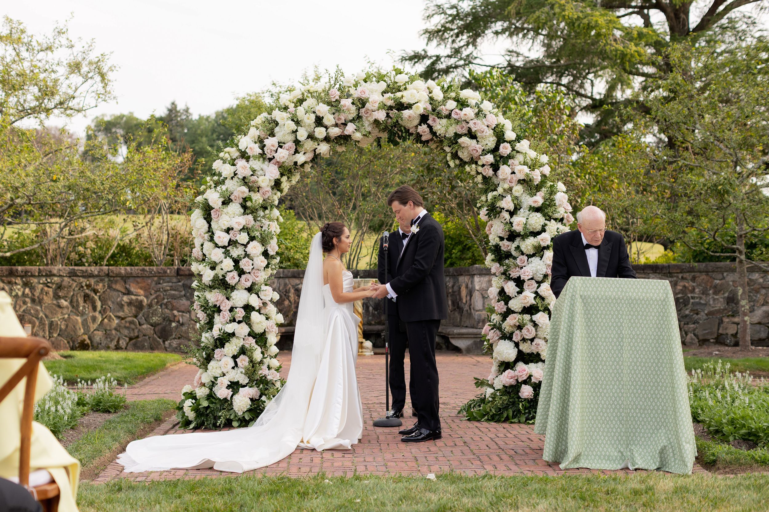An outdoor wedding ceremony setup with a white floral arch and garden setting.