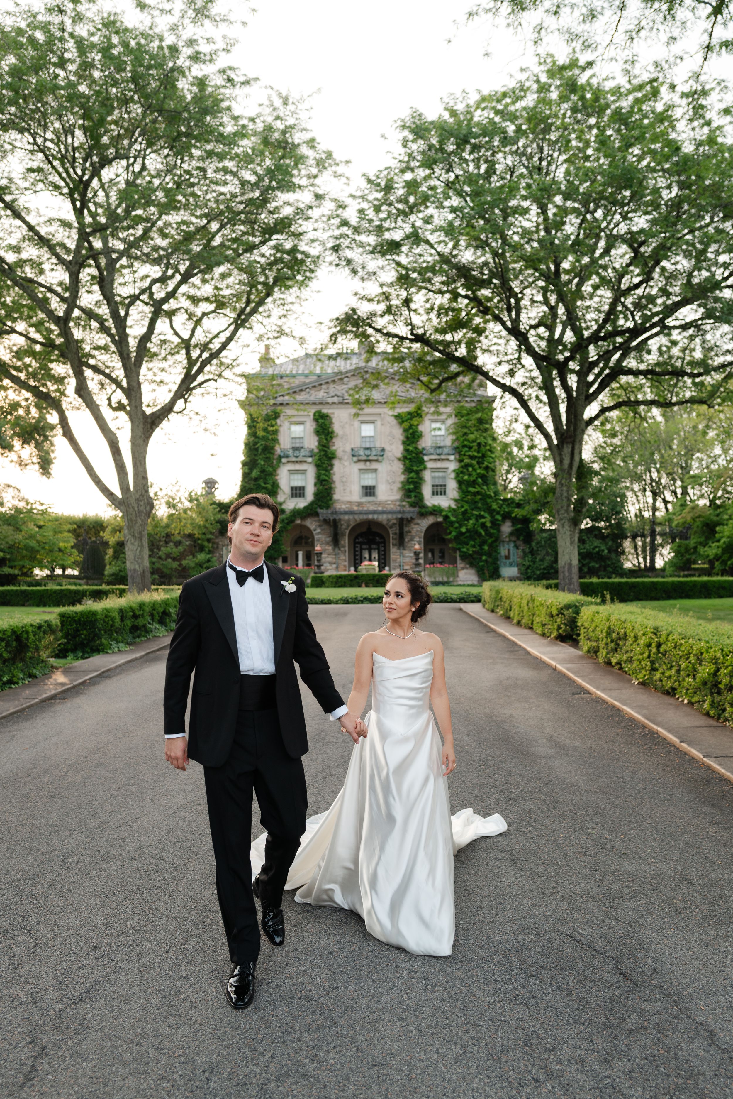 A couple in wedding attire walks and dances along a tree-lined driveway at their elegant outdoor venue.