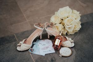 Wedding accessories including white bridal shoes, bouquet of cream roses, and gift tag arranged on stone floor.