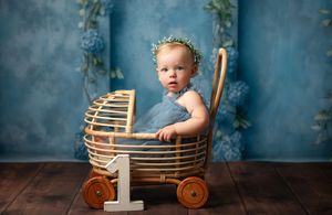 Baby with floral crown sits in vintage wooden wagon cart against blue backdrop with number one decoration.