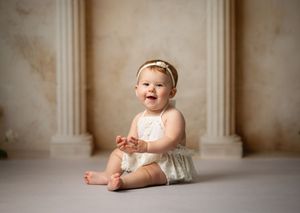 A happy baby wearing a headband and light colored outfit sits on the floor in a studio with elegant columns.