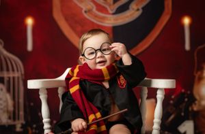 Small child wearing a Hogwarts uniform and glasses sits in a white chair with red and gold decor and candles in background.
