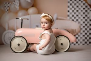 A small child in a white dress sits beside a vintage-style pink toy car in a softly lit nursery setting.