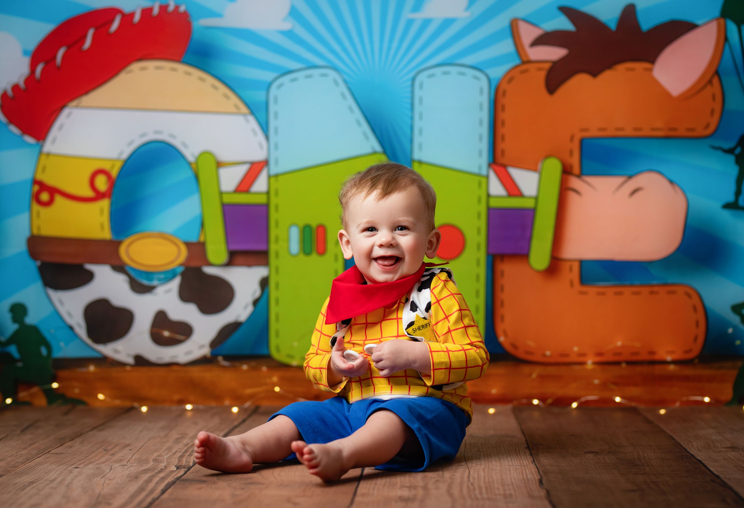 A young child sits on wooden flooring against a colorful toy train backdrop while wearing a yellow and red outfit.