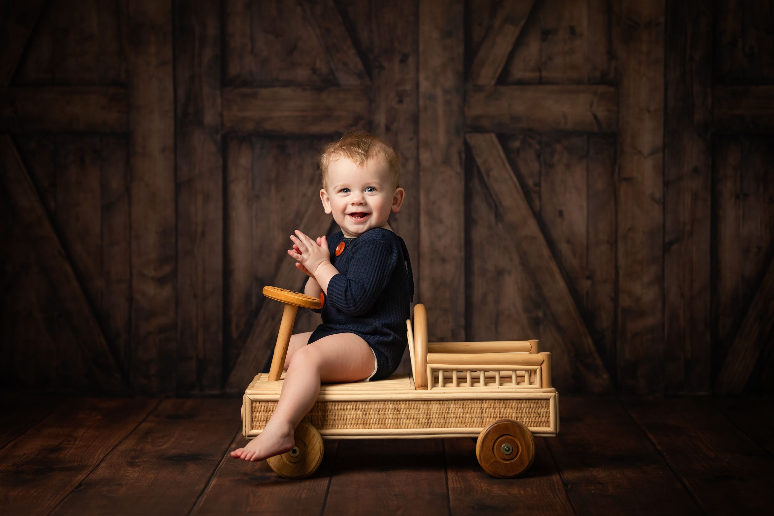 Baby wearing navy romper sits in wooden vintage toy car against rustic barn door background.