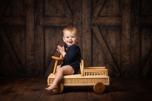Baby wearing navy romper sits in wooden vintage toy car against rustic barn door background.