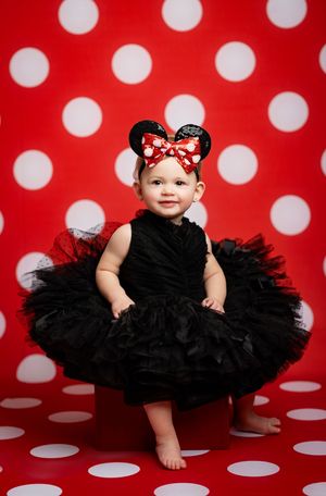 Baby in black tutu dress and red polka dot bow sits against red and white polka dot backdrop in photo series.