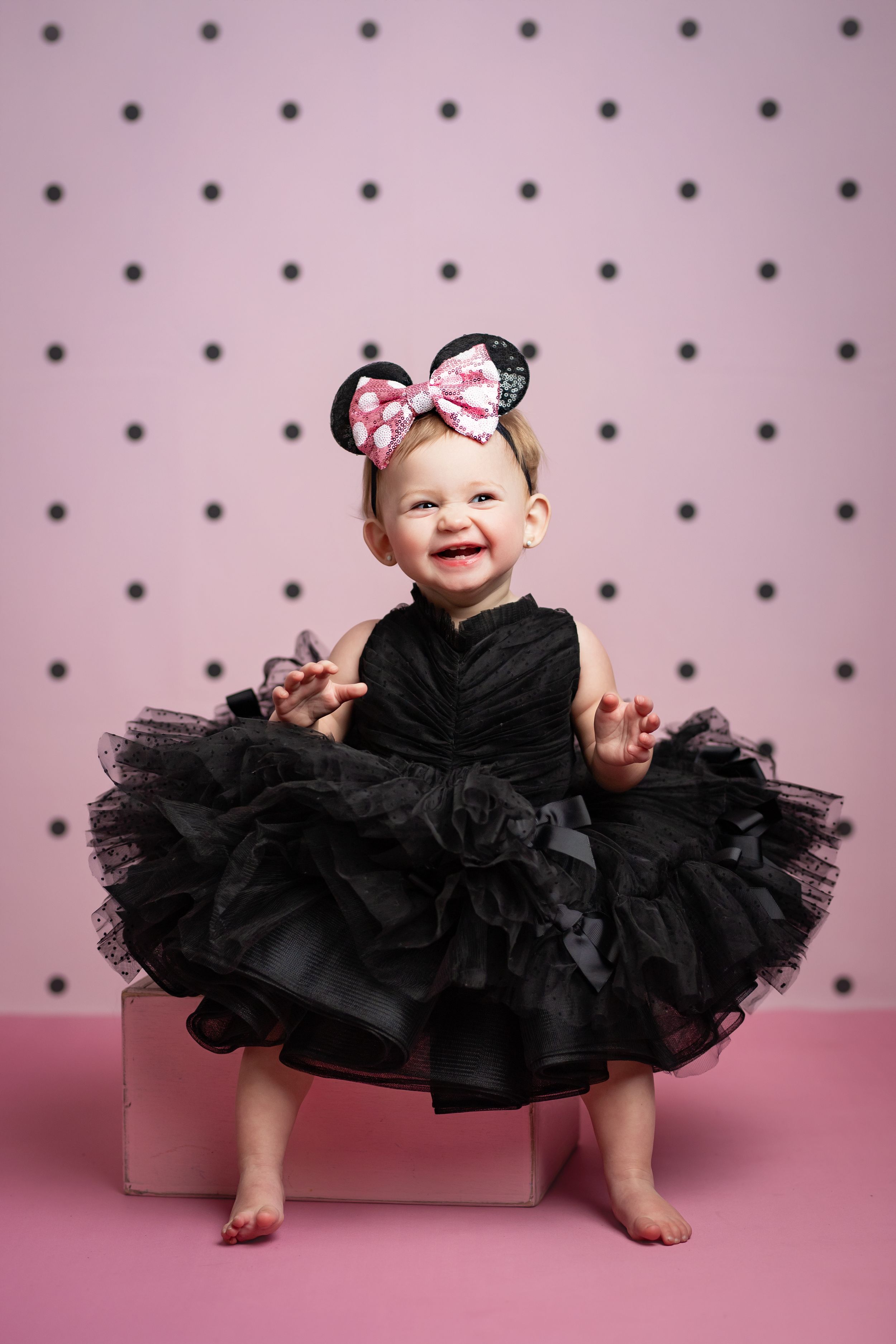 A baby wearing a black tutu dress and bow sits on a pink backdrop with polka dots in three different poses.
